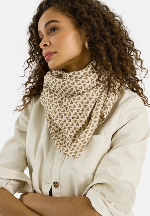 Mujer con cabello rizado que lleva aretes dorados, una chaqueta beige y una bufanda beige con un patrón de corazones marrones, mirando hacia un lado.