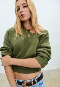 Jonge vrouw met blond haar die een olijfgroene off-shoulder sweater, kraalkettingen, lichtblauwe jeans en een bruine riem met een decoratieve gesp draagt.