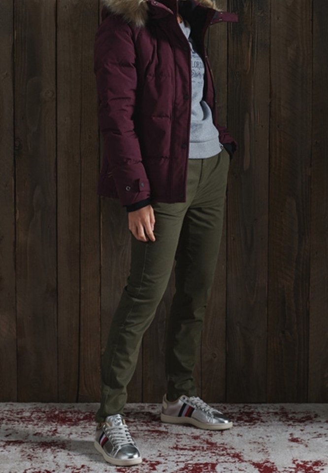 Personne portant une veste bordeaux à capuche en fourrure, un sweat-shirt gris clair, un pantalon vert olive et des baskets argentées métalliques, debout sur un tapis rouge et blanc.