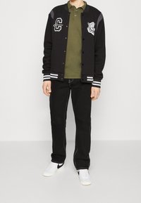 Svart varsityjacka med vita ränder på ärmsluten, grön piké, svarta jeans och vita sneakers med svarta accenter.