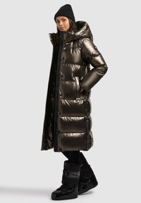 Hosszú, puffadt kabát fémes bronzszínben, kapucnival, elöl cipzárral és oldalzsebekkel. Fekete bélelt csizmával és kötött sapkával párosítva.