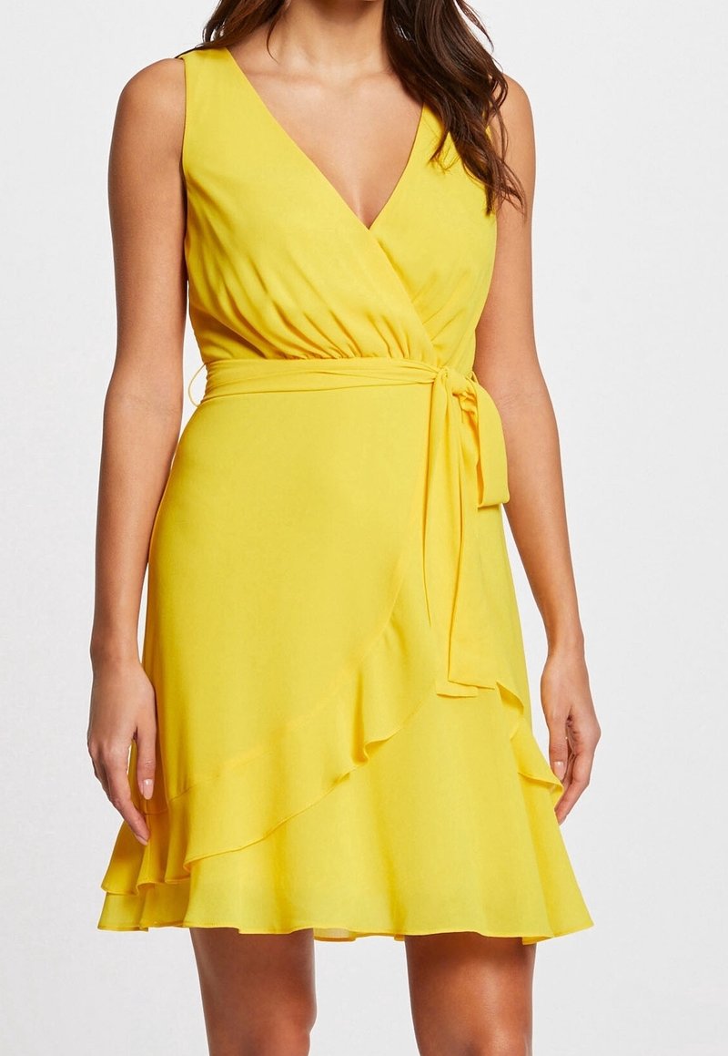 Femme portant une robe portefeuille jaune sans manches avec col en V, taille ceinturée et ourlet volanté, debout devant un fond uni.