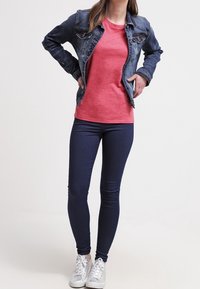 Giacca di jeans sopra una maglietta a righe rosse e bianche, abbinata a jeans skinny blu scuro e sneaker argento. Outfit casual in sovrapposizione.