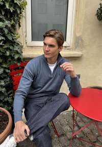 Jaunas vyras mėlynu megztiniu su užtrauktuku ir džinsais sėdi sukryžiavęs kojas raudonoje kėdėje prie raudono stalo šalia lango ir išorinės sienos, apaugusios erškėčiais.