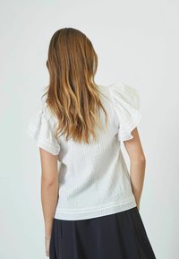 Blusa bianca in cotone con tessuto texturizzato, maniche a sbuffo e dettagli in pizzo all'orlo. Indossata con una gonna scura, vista da dietro.