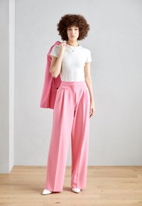 Pink high-waisted trousers with pleats paired with a white fitted t-shirt. Model holds a matching pink blazer. The setting is neutral-colored.