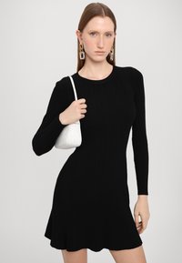 Zwarte geribbelde mini-jurk met lange mouwen en een uitlopende zoom, gecombineerd met een kleine witte handtas met textuur. Grote geometrische oorbellen.
