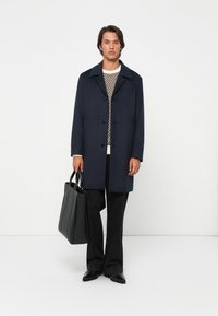 Uomo che indossa un cappotto blu scuro, un maglione a fantasie, pantaloni neri e scarpe, mentre tiene una borsa a tracolla in pelle nera, in piedi davanti a uno sfondo bianco.