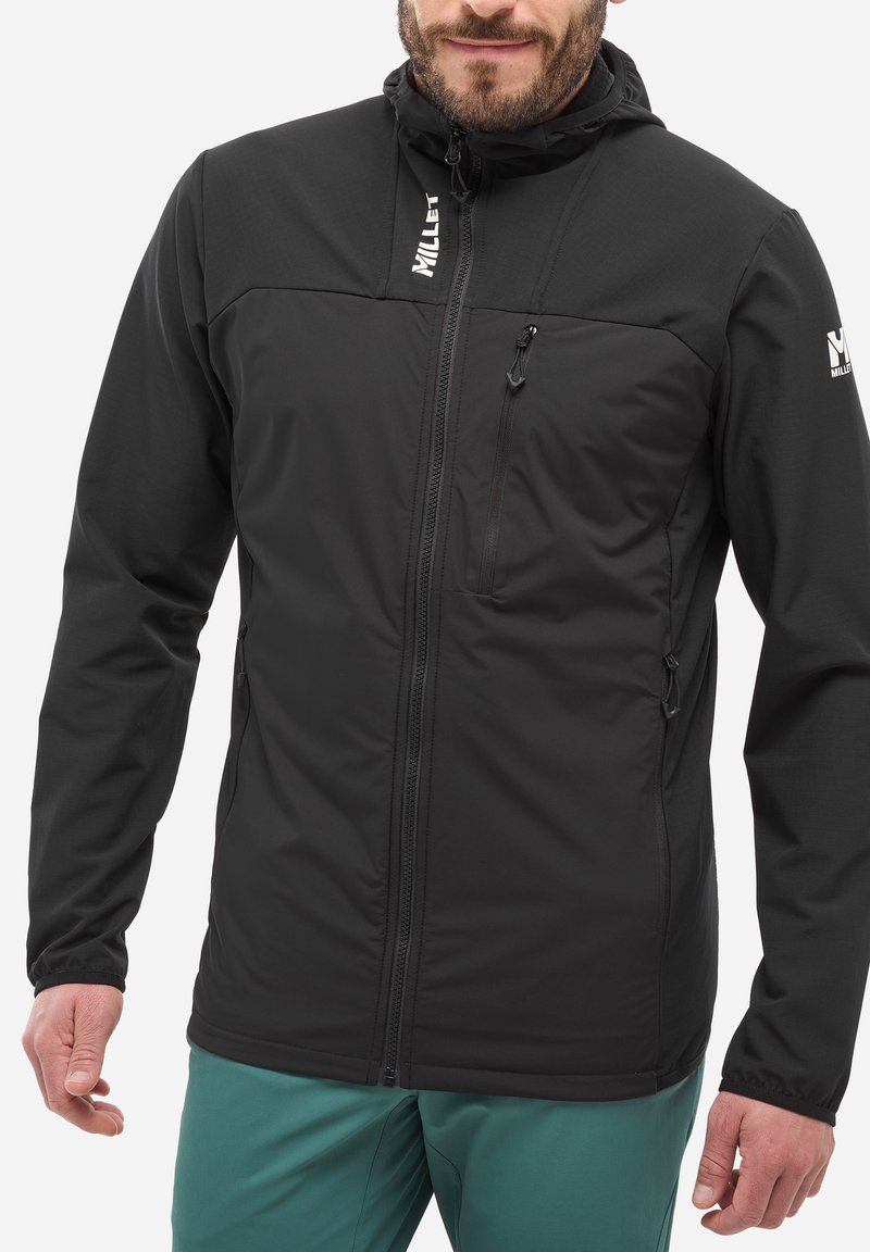 Homme portant une veste noire zippée avec des poches zippées sur la poitrine et sur les côtés, et un pantalon bleu-vert, se tenant debout avec les mains détendues sur les côtés.
