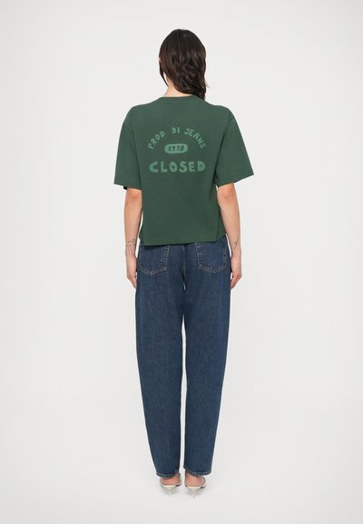 Πράσινο βαμβακερό T-shirt με χαλαρή εφαρμογή, με το κείμενο "CLOSED" και το λογότυπο στην πλάτη, συνδυασμένο με φαρδιά μπλε τζιν παντελόνια.