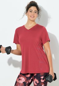 Czerwona koszulka sportowa z dekoltem w serek i krótkimi rękawami, zestawiona z żywymi, kwiecistymi legginsami. Modelka trzyma czarną hantlę w jednej ręce.