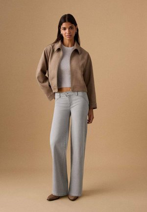 Chaqueta de ante marrón recortada, top gris ajustado con textura acanalada, vaqueros de pierna ancha en gris claro y zapatos marrones sobre un fondo neutro.