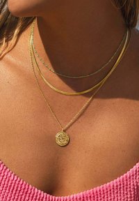 Collares de oro en capas sobre la piel; presenta una cadena gruesa, una cadena delicada con acentos azules y un colgante con un diseño detallado.