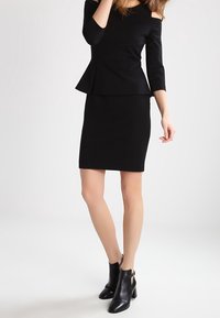 Schwarzes Peplum-Kleid mit schulterfreien Ärmeln, taillierter Silhouette und knielanger Rocklänge. Kombiniert mit schwarzen Knöchelstiefeln.