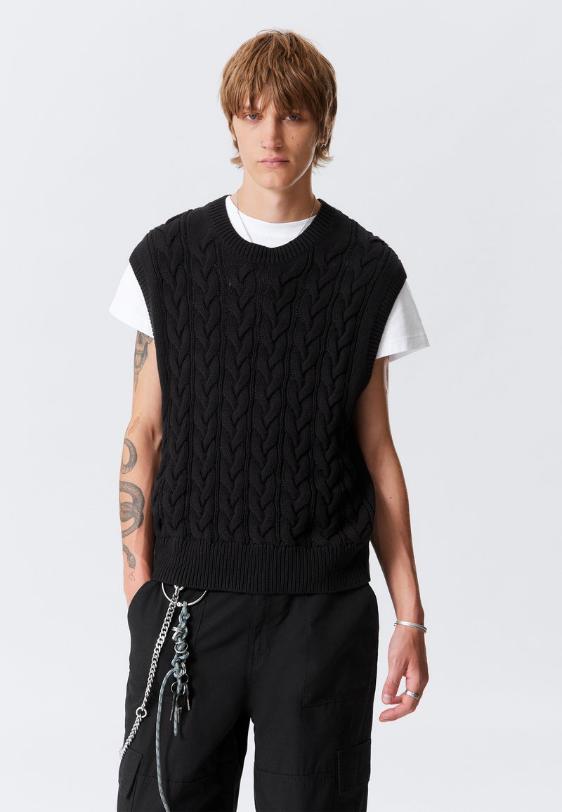 Fekete kötött ujjatlan pulóver kábelmintával, bordázott nyakkal és szegéllyel. Fehér póló fölé viselve, fekete nadrággal, amelyen fém kiegészítők vannak.