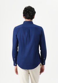 Camisa de manga larga azul marino hecha de tejido suave, con cuello abotonado y dobladillo curvado, con costuras visibles y botones en los puños.