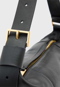 Curea de geantă din piele neagră cu cataramă metalică aurie. Prezintă găuri ajustabile și o textură netedă. Fermuar vizibil pe marginea genții.