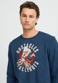 Tamno plavi dukserica od pamuka s okruglim izrezom, s grafičkim otiskom lika koji vozi bicikl uz tekst "PREPREKE SU IZAZOVI."