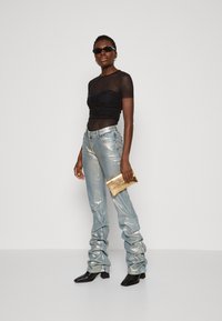 Top negro transparente con detalles fruncidos, jeans metálicos azul claro, cartera dorada y botas negras de tacón. Los accesorios incluyen gafas de sol y aretes.