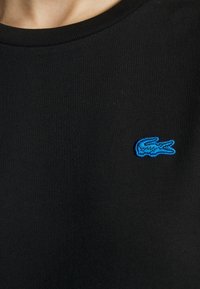 Camiseta negra de cuello redondo, hecha de una tela suave, presenta un pequeño logo de cocodrilo bordado en azul en el pecho.