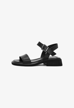 Sandaña de cuero negro con punta abierta, correa ajustable en el tobillo y tacón bajo. Textura suave con un diseño minimalista.