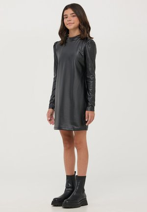 Czarna sukienka z sztucznej skóry z długim rękawem i wysokim dekoltem, o delikatnym połysku, sięgająca do połowy uda, zestawiona z czarnymi botkami za kostkę.