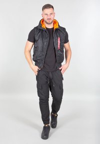 Vīrietis, kurš dodas uz priekšu, ģērbts melnās cargo biksēs, melnā T-kreklā, melnā vestē ar oranžu iekšējo oderi un melnās sporty kedās.