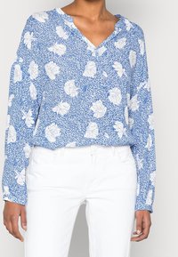 Blusa floreale blu con motivi di rose bianche, maniche lunghe e scollatura a V. Indossata con pantaloni bianchi. Tessuto leggero, vestibilità comoda.