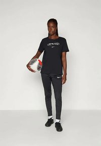Čierne tričko Nike s logom "SPURS", skombinované s čiernymi legínami so vzorom. Model drží viacfarebnú futbalovú loptu a má na sebe čierne tenisky.