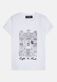 Vit bomull t-shirt med svart linjekonst av ett café, med texten "Café de Karl" under illustrationen, korta ärmar och rund halsringning.