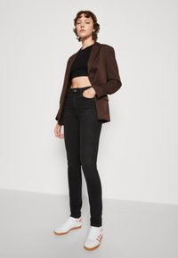 Smeđi sako preko crnog kratkog topa, uparen s crnim trapericama visokog struka i bijelim tenisicama s narančastim naglascima. Glatke teksture, krojeni fit.