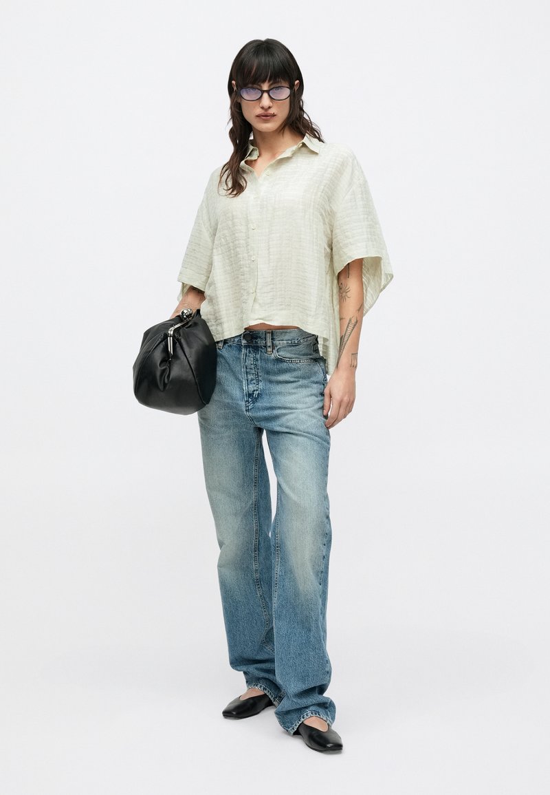 Lysegrøn kortærmet skjorte med knapper, lavet af tekstureret stof; kombineret med falmede blå jeans og en sort håndtaske; modellen bærer briller.