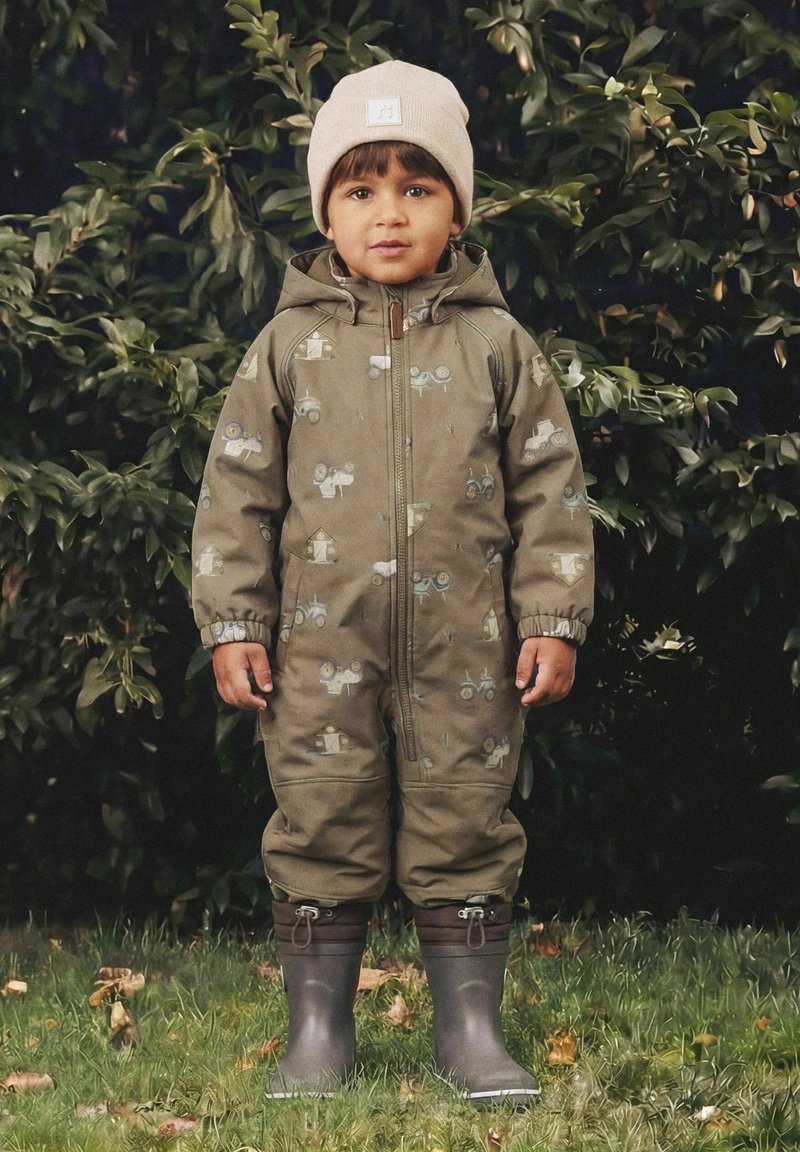 Jeune enfant se tenant à l'extérieur portant une combinaison verte à motifs, des bottes en caoutchouc grises et un bonnet beige en tricot devant un fond feuillu.
