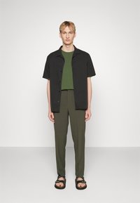 Camisa preta de manga curta, t-shirt verde por baixo, calças verde-oliva e sandálias negras com tiras. Simples, com um corte ajustado.