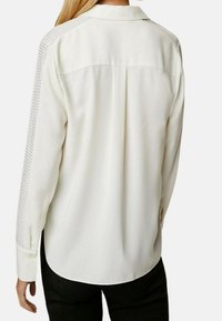 Camicia bianca con colletto, maniche lunghe e dettagli perforati sulle spalle. Tessuto morbido con orlo arrotondato e polsini con bottone.