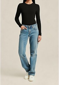 Personne portant un haut noir ajusté à manches longues, un jean droit bleu clair avec une main dans la poche, et des baskets blanches, debout.