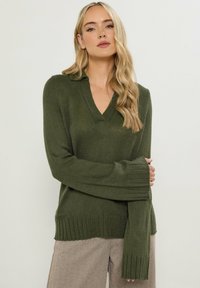Sweter dzianinowy w kolorze ciemnozielonym z dekoltem w serek, z prążkowanymi akcentami przy mankietach i dolnym wykończeniu, wykonany z materiału o fakturze. W komplecie z jasnymi spodniami w wzory.