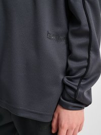 Zbliżenie osoby noszącej ciemnoszarą bluzę z długim rękawem hummel Performance oraz pasujące spodnie, z widocznym ramieniem i dłonią.