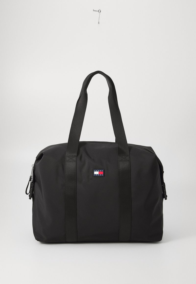 Crni torba s dvostrukim materijalnim ručkama, zatvaranjem na zip i dekorativnom oznakom logotipa u crvenoj, bijeloj i plavoj boji na prednjoj strani. Glatka tekstura.
