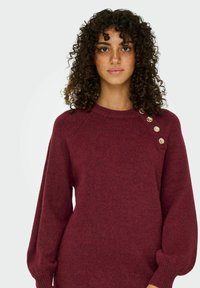Pull bordeaux en tricot doux, avec un col rond, des manches surdimensionnées et des boutons décoratifs dorés sur l'épaule gauche.