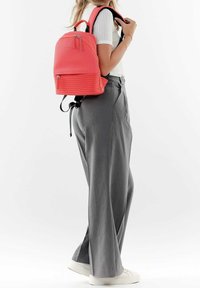 Zaino color corallo con trama a coste orizzontali, doppia cerniera e spallacci regolabili; abbinato a pantaloni grigi a gamba larga e sneaker bianche.