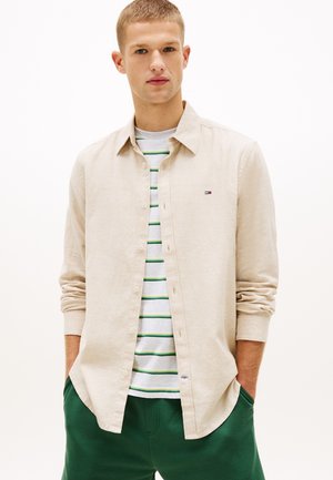 Joven vestido con camisa beige abotonada sobre camiseta blanca con rayas verdes, pantalones verdes, de pie con las manos en los bolsillos.