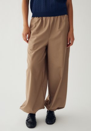 Persona con pantalones holgados color canela, una camiseta sin mangas azul oscuro y zapatillas negras sin cordones, de pie sobre un fondo blanco.