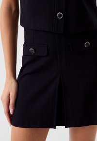 Schwarzes maßgeschneidertes Kleid mit tailliertem Oberteil, einem Frontschlitz und zwei Brusttaschen mit runden schwarzen Knöpfen mit glänzenden Akzenten.