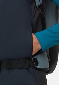 Schwarzer Rucksack mit grauen Akzenten; die Hand des Benutzers greift in eine Seitentasche. Verstellbarer Gurt sichtbar, mit einer robusten Schnalle.