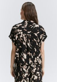 Blusa a maniche corte con una base nera decorata da motivi astratti beige e marroni, dotata di colletto e una vestibilità ampia.