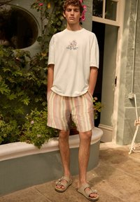 Camiseta oversized verde claro con estampado floral, pantalones cortos a rayas en tonos pastel y sandalias gris claro. Ambiente al aire libre con vegetación.
