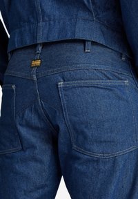 Donkerblauwe denim jeans met een gladde textuur, contrasterende stiksels en een zichtbaar merklabel op de achterste tailleband. Achteraanzicht met zakken.