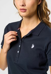 Marineblå ribbestrikket polo med treknappslukking, hvit logo broderi på brystet og kontrasterende innerkrage. 
