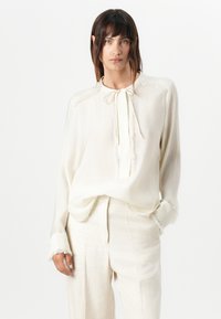 Lys krembluse med knyting ved halsen, teksturert detaljer langs yoken og lett frynsete mansjetter, kombinert med lyse beige linbukser.